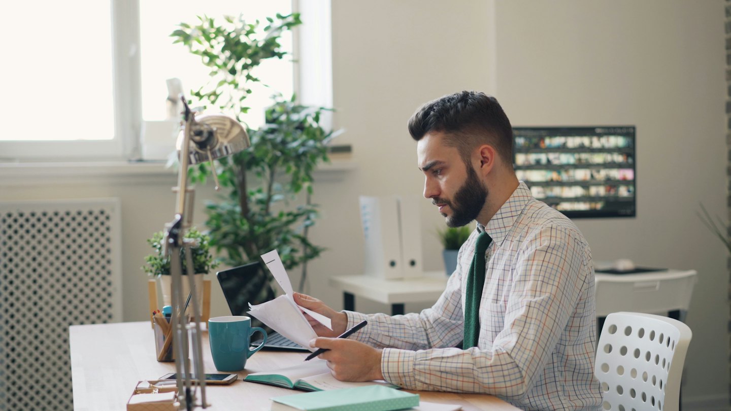 Man in plaid shirt and green tie reviews documents at desk.